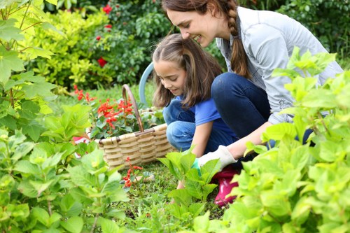 A woman and a young girl are crouched in a lush, well-maintained garden, engaging in outdoor gardening activities. They are surrounded by vibrant green foliage, including a variety of shrubs and leafy plants, with a few flowering bushes displaying small red flowers. The garden features a neat, striped lawn with dense grass, bordered by flower beds and planting borders that create a structured landscape. In the background, there are mature trees providing shade and natural backdrop, with sunlight filtering through the leaves, suggesting a clear day with bright weather. The woman appears to be guiding or assisting the girl as they reach towards the plants, possibly planting or watering, while the girl holds a small wicker basket, indicating active gardening work. The scene depicts a peaceful, well-kept outdoor space that highlights gardening and outdoor maintenance, fitting for a professional gardening service in the local area serviced by Hedge Trimming Archway, near the postcode for the town of Archway.