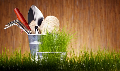 A close-up of a gardening scene featuring a small metal bucket filled with various garden tools including a trowel, a hand rake, and a pair of pruning scissors, with a ball of natural twine resting on top of bright green grass. The tools have red and silver handles, contrasting with the lush, vibrant grass and the weathered, wooden surface background, suggesting outdoor gardening activity. The scene emphasizes gardening maintenance elements and outdoor space preparation, typical of services offered by Hedge Trimming Archway in the UK, with natural tones and practical equipment suitable for lawn and garden care in local gardens or landscaped yards.